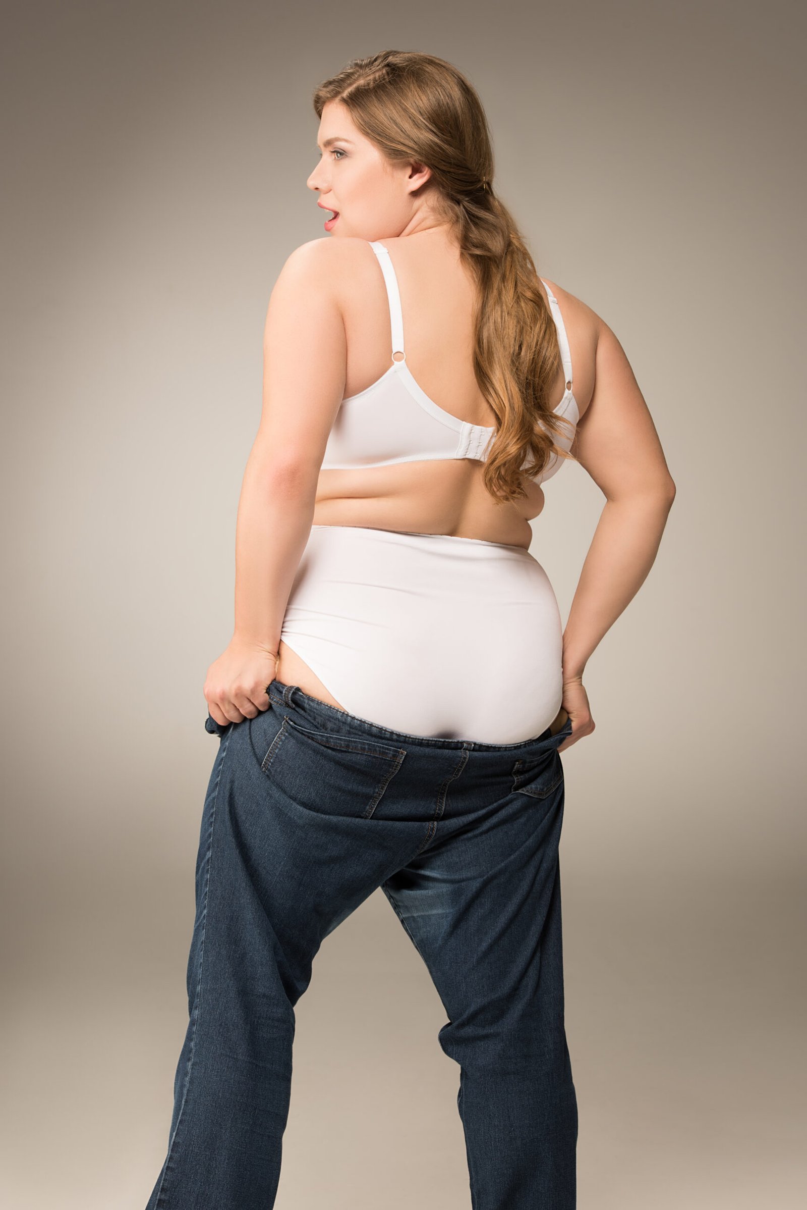 Overweight girl in white underwear wearing big jeans, isolated on grey